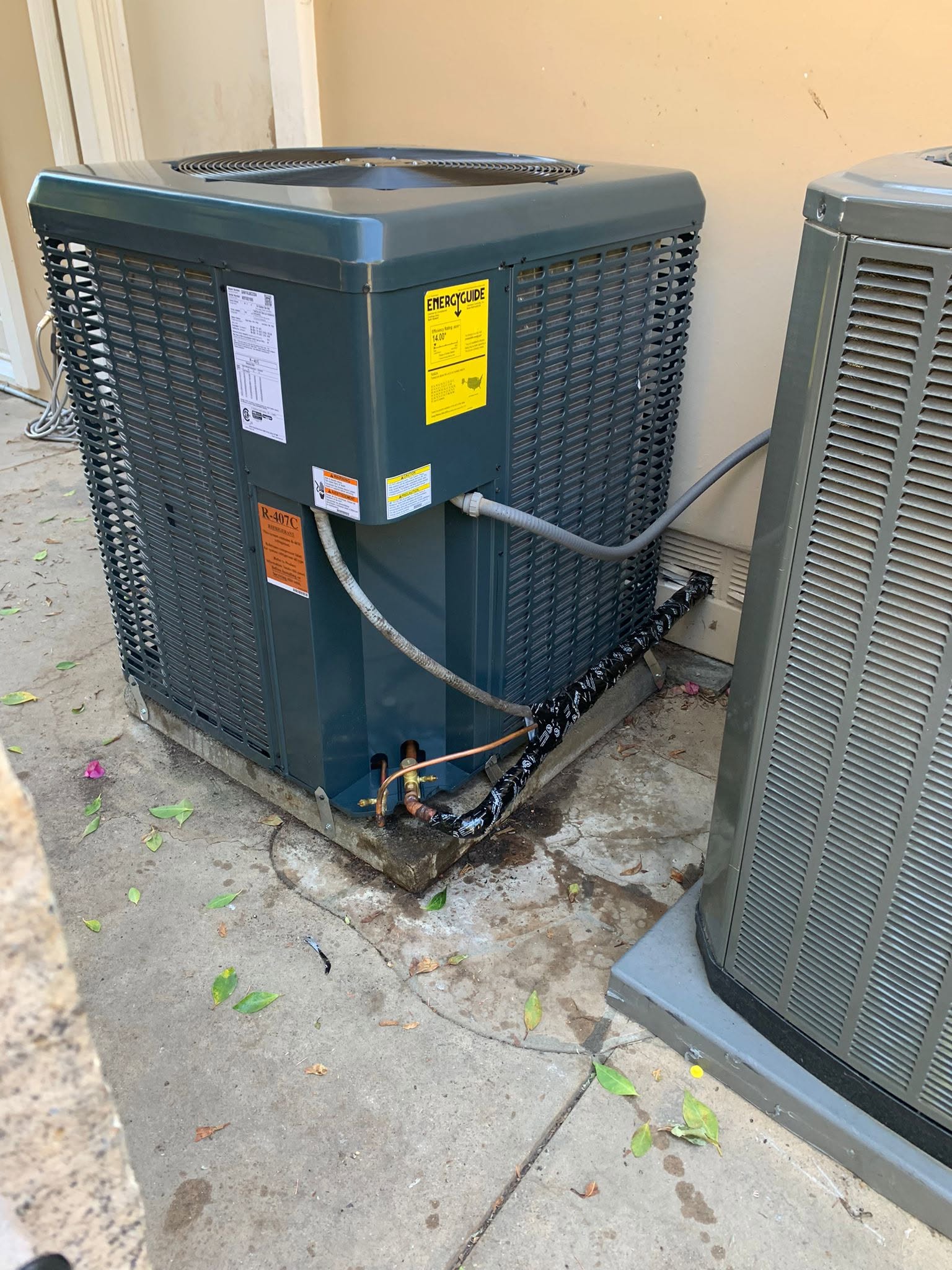 Outdoor HVAC unit with visible wiring and pipes, some water leakage on the concrete below; leaves and debris scattered around the base.