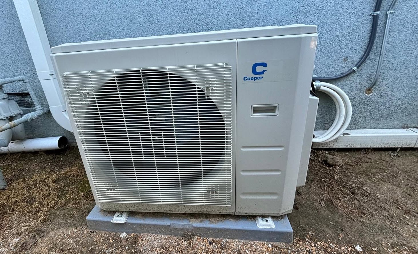 A Cooper outdoor air conditioning unit is installed on a concrete pad next to a blue wall, with pipes connected to the building.