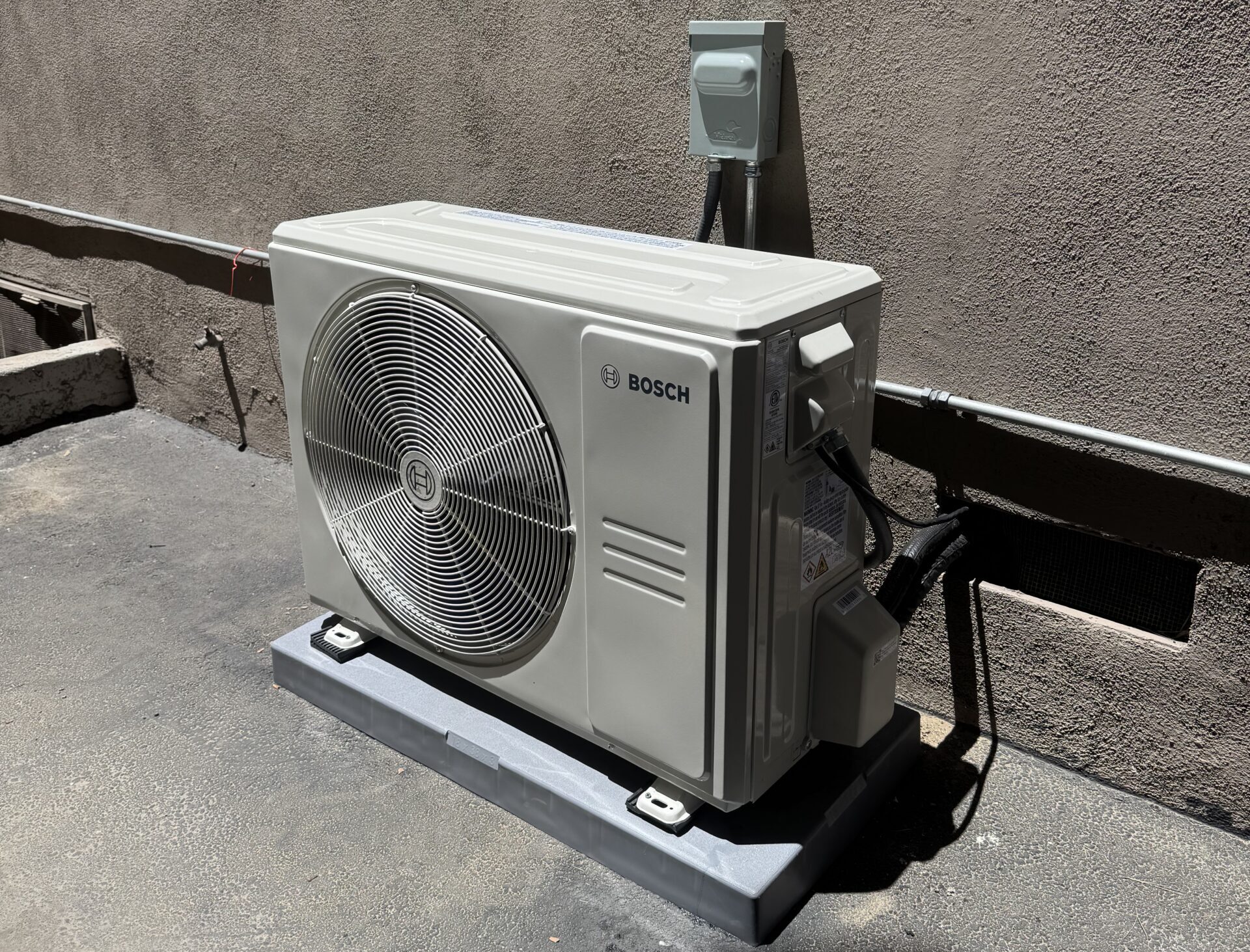 A Bosch outdoor air conditioning unit is installed on a concrete pad against a stucco wall, with power and refrigerant lines connected.