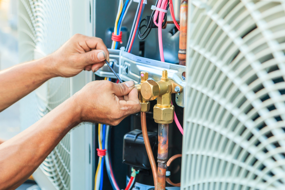 A person uses a wrench to adjust valves and wiring on an outdoor air conditioning unit.