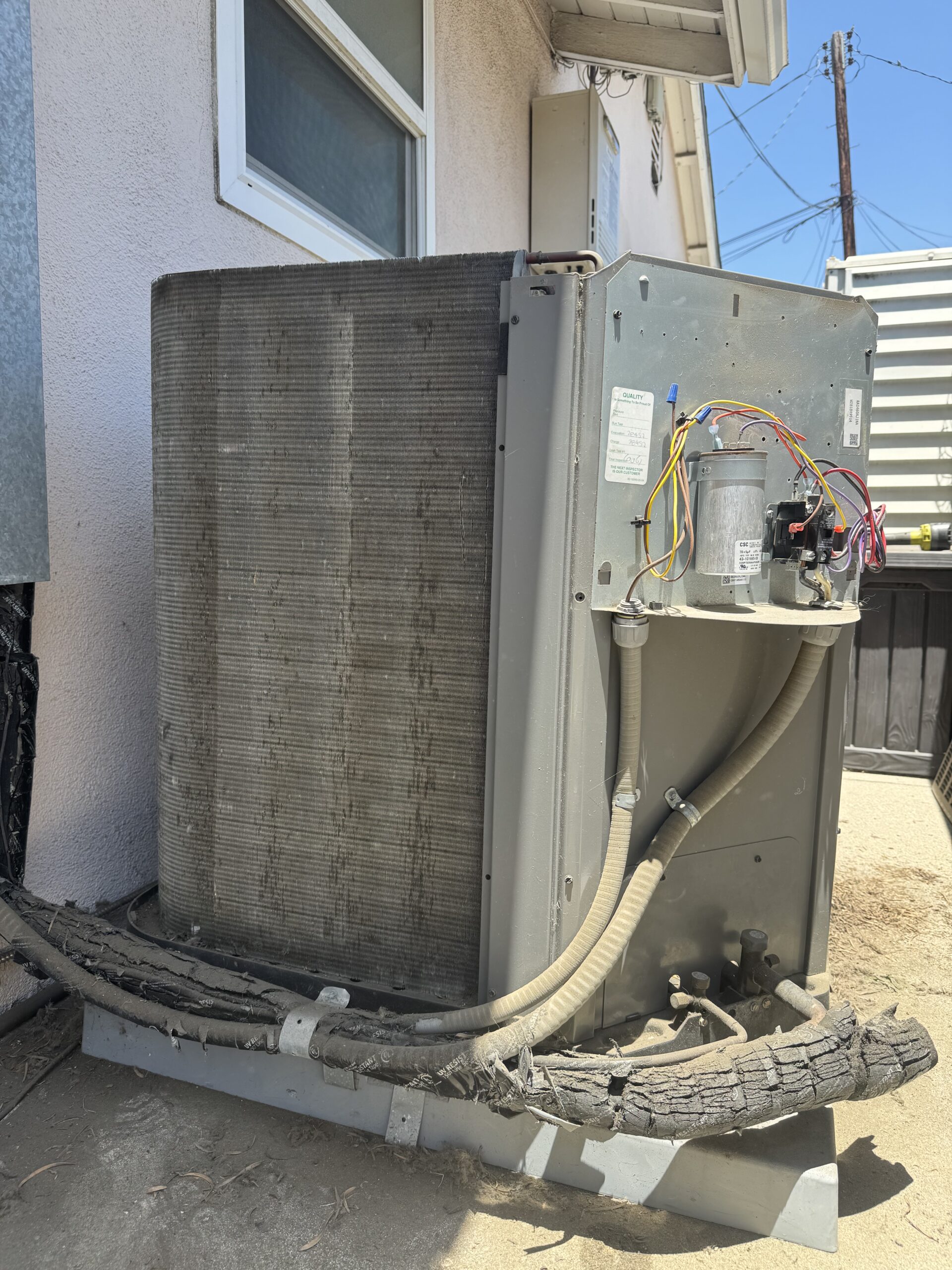 Outdoor air conditioning unit with exposed wiring and damaged insulation, positioned next to a building wall on a concrete pad.
