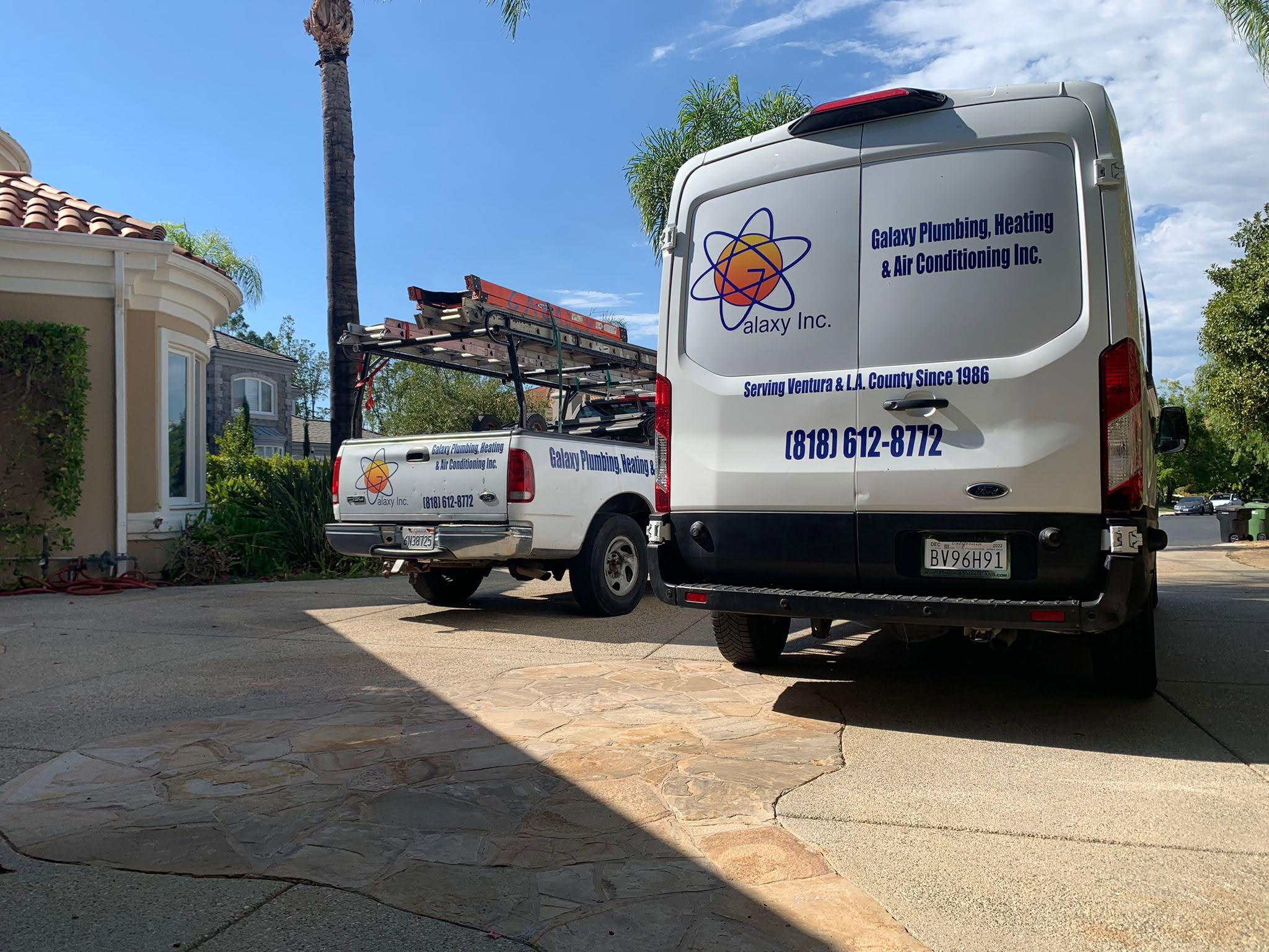 Two Galaxy Inc. service vehicles are parked in a sunny residential driveway. Text on the vans advertises plumbing, heating, and air conditioning services and includes a phone number.
