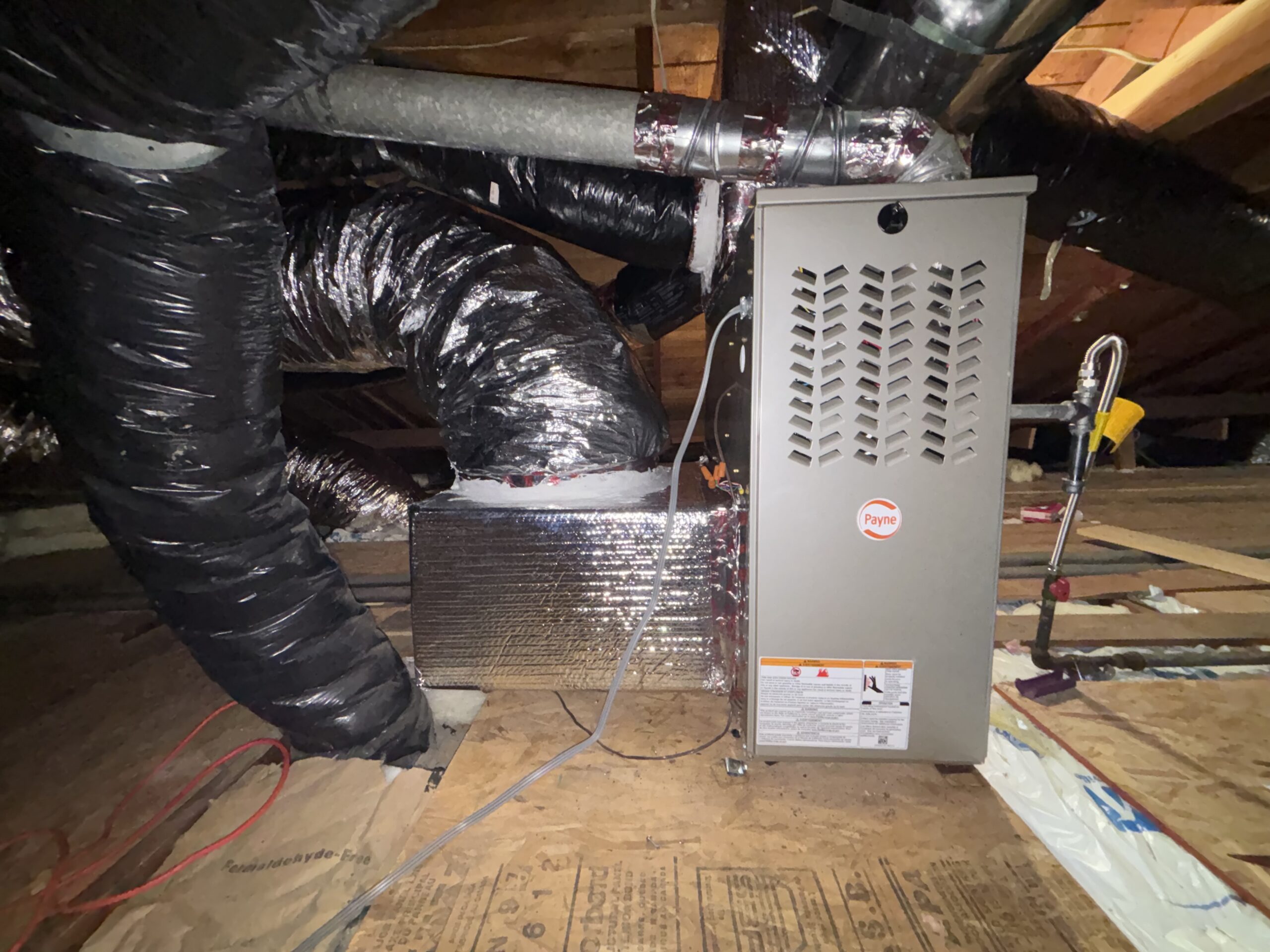 A furnace unit with ductwork and insulation is installed in an attic with exposed wooden beams and wiring visible on the floor.