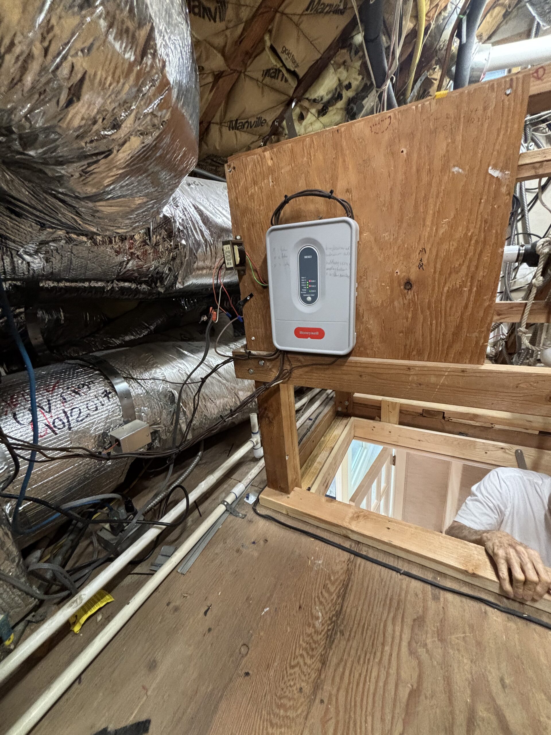 An HVAC system with insulated ducts, wiring, and a control box is installed in an attic. A person's arm is visible near a wooden opening in the floor.