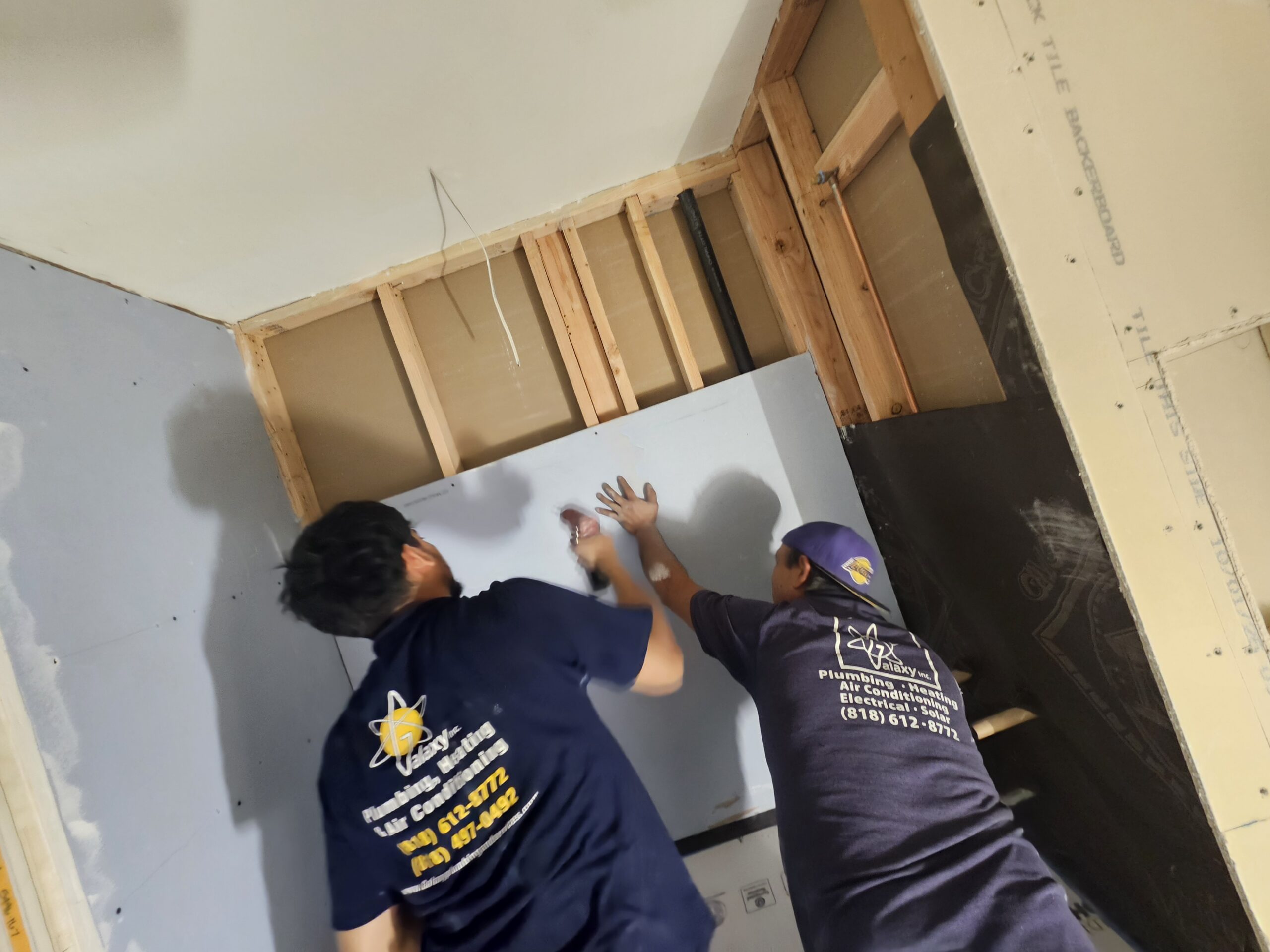 Two people install a drywall sheet on a wall frame in a partially constructed room. Both wear blue shirts with yellow logos and text on the back.