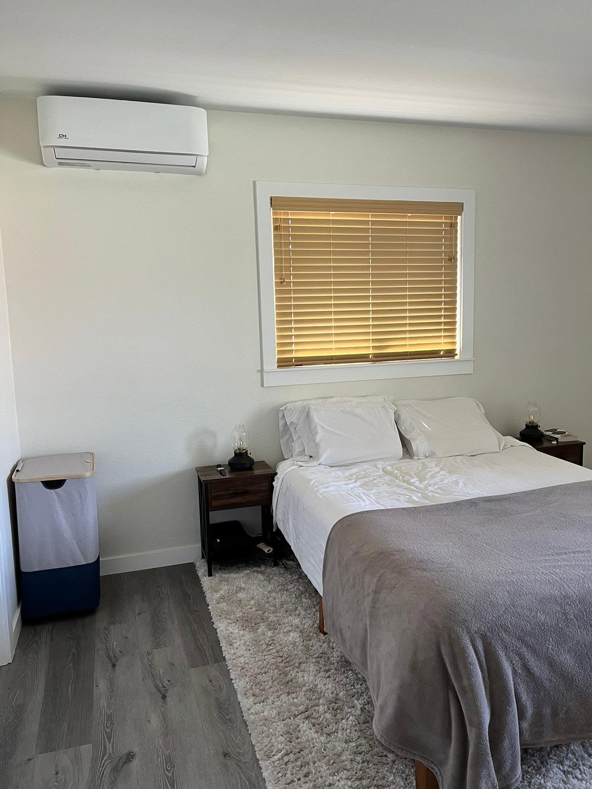 A neatly made bed with white sheets and a gray blanket is between two nightstands. A window with closed blinds is above the bed, and an air conditioner and trash bin are on the left wall.