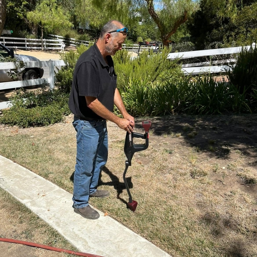 A man stands on a lawn using a metal detector, with a white fence and greenery in the background on a sunny day.