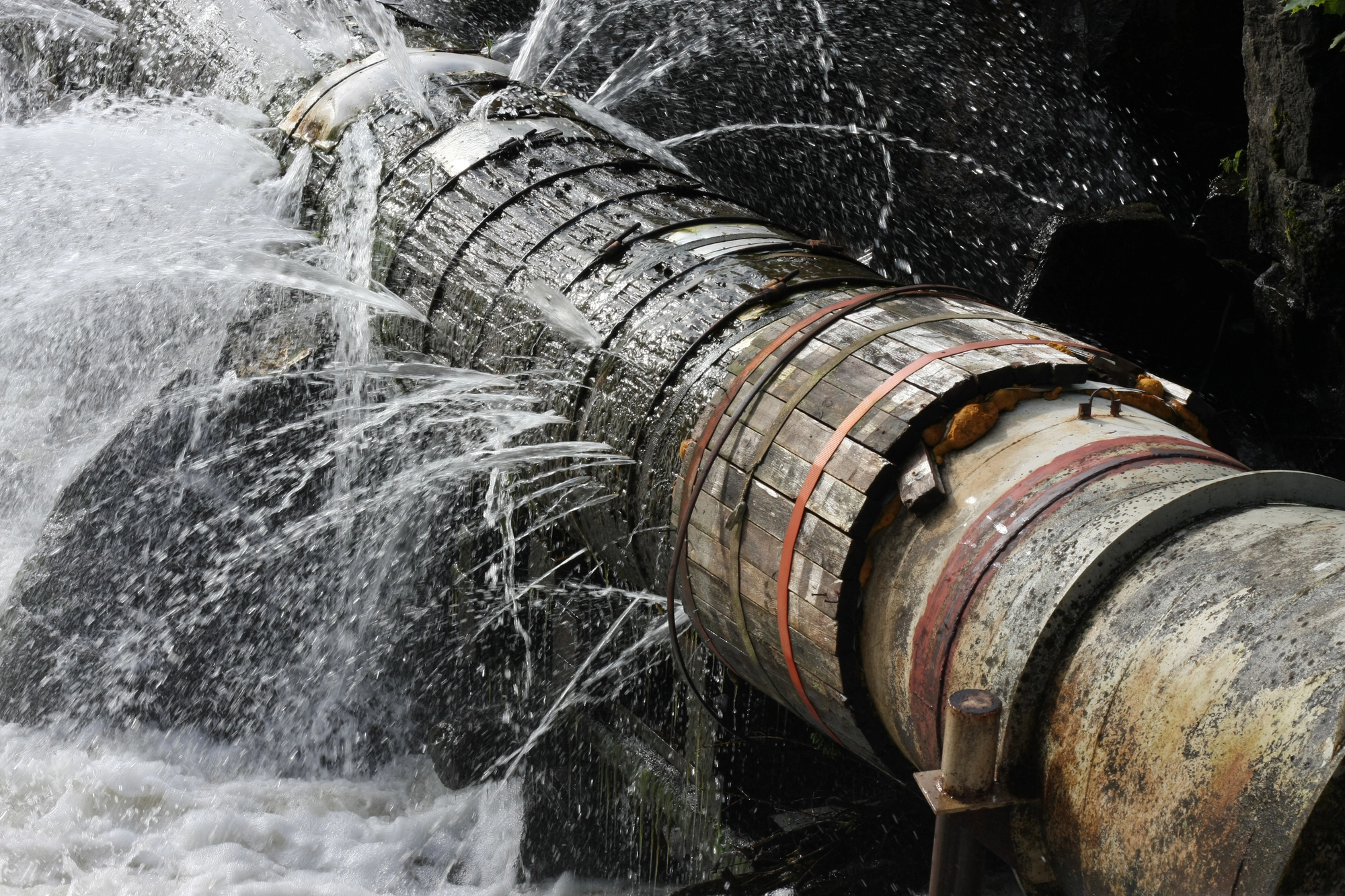Large metal pipe leaking water from multiple cracks and joints, with water spraying out forcefully against a dark background.