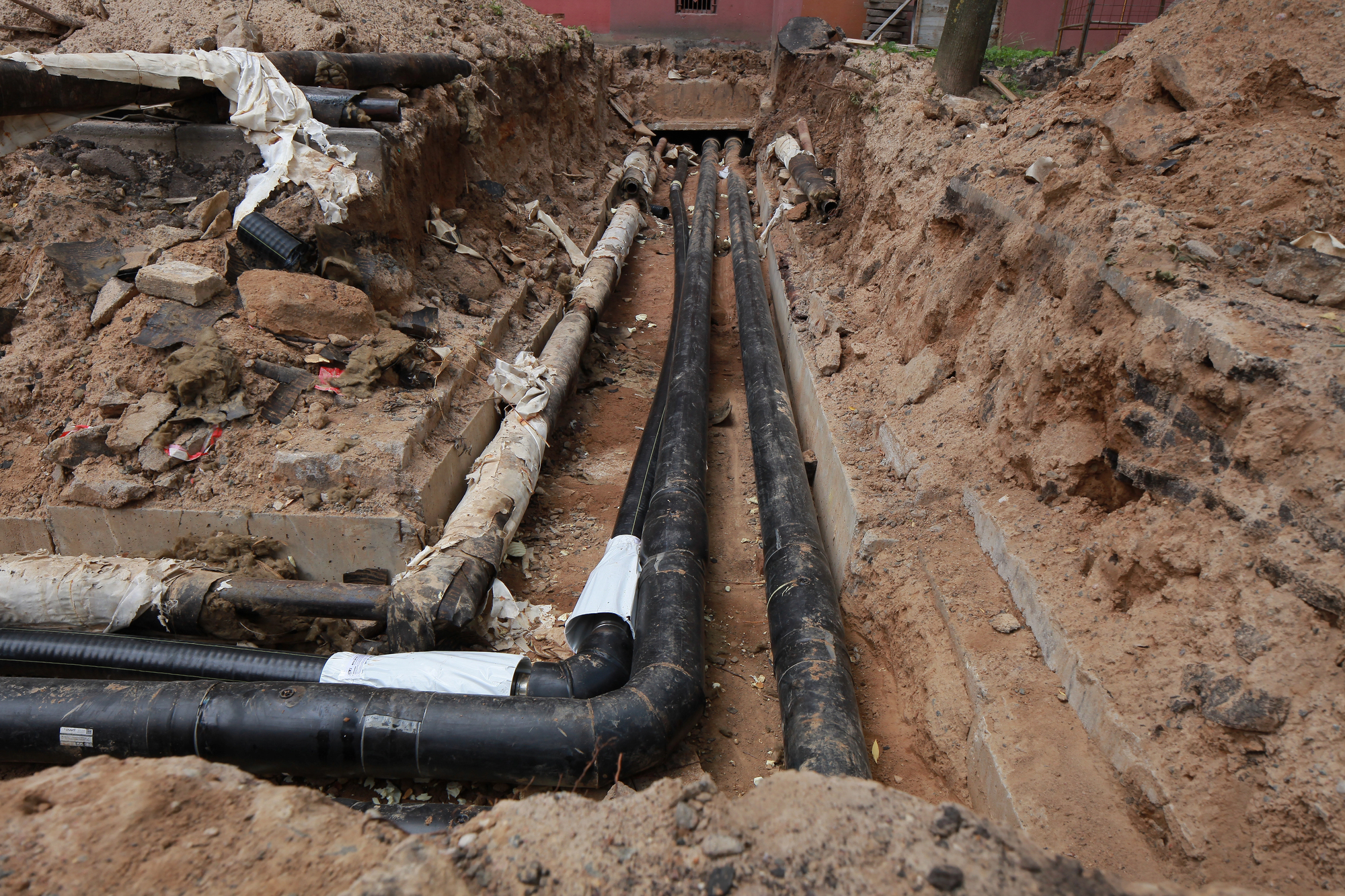 Excavated trench with multiple large black pipes running underground, surrounded by dirt and construction debris.