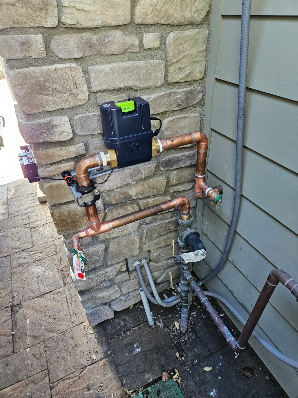 Outdoor plumbing system with copper pipes, a black control unit, valves with tags, and multiple connections attached to a stone and siding exterior wall.
