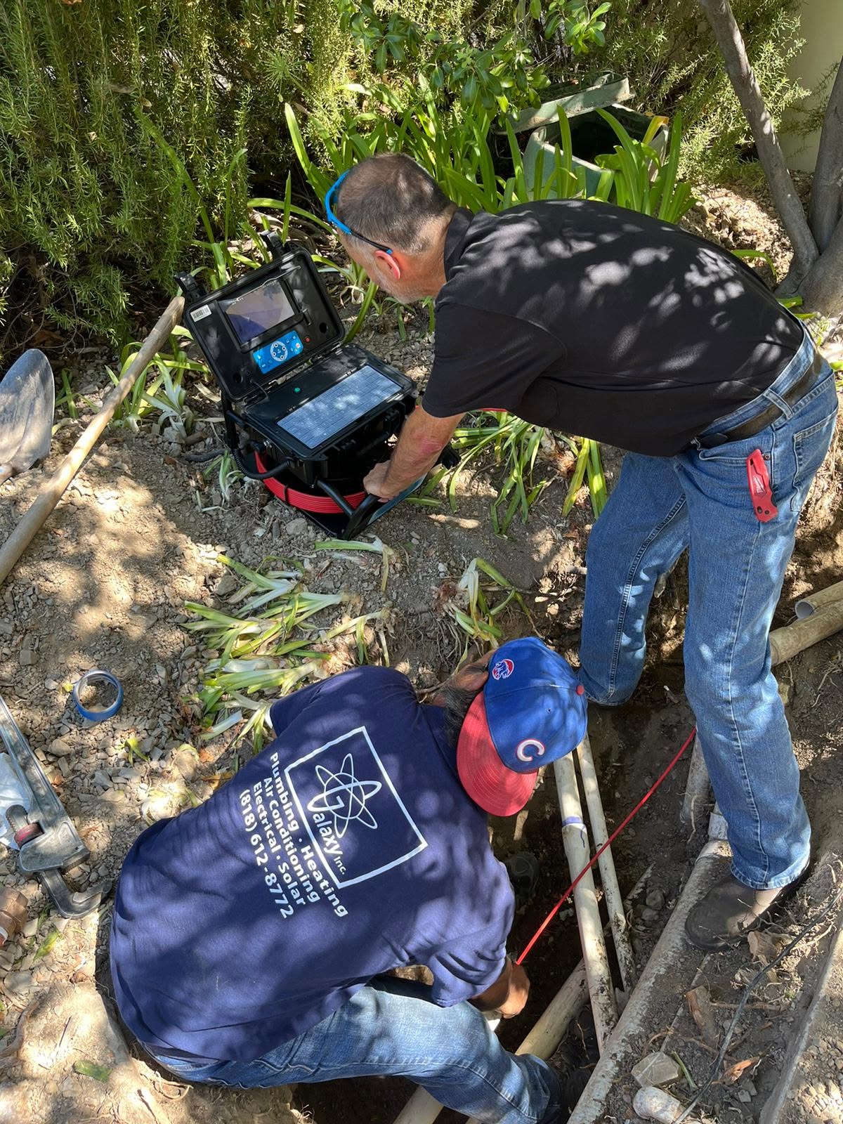 Two men inspect underground pipes outdoors using a camera system; one monitors the screen while the other works near the exposed piping.