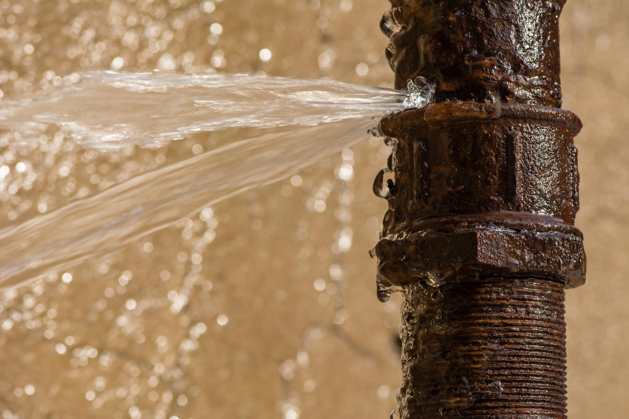 A rusty metal pipe leaking water from a crack, with water spraying out forcefully to the left side of the image.