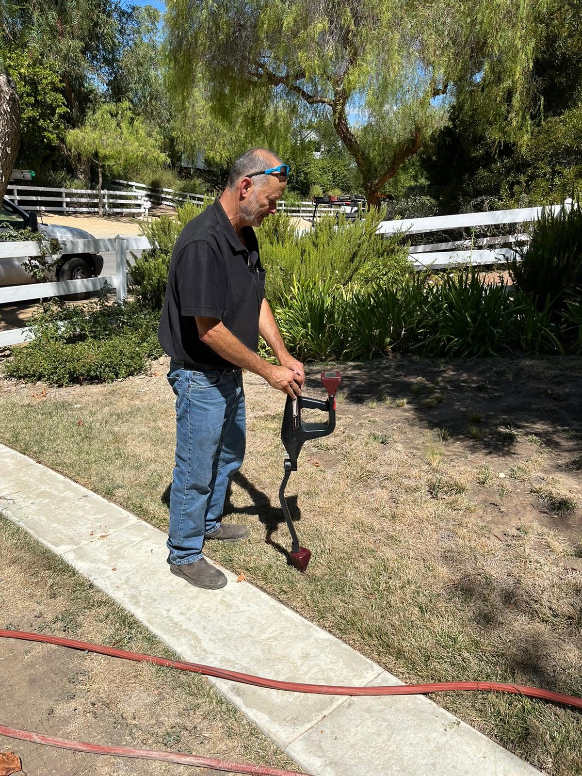 A man in sunglasses and a black shirt uses a metal detector on a grassy area next to a sidewalk, with trees and a white fence in the background.
