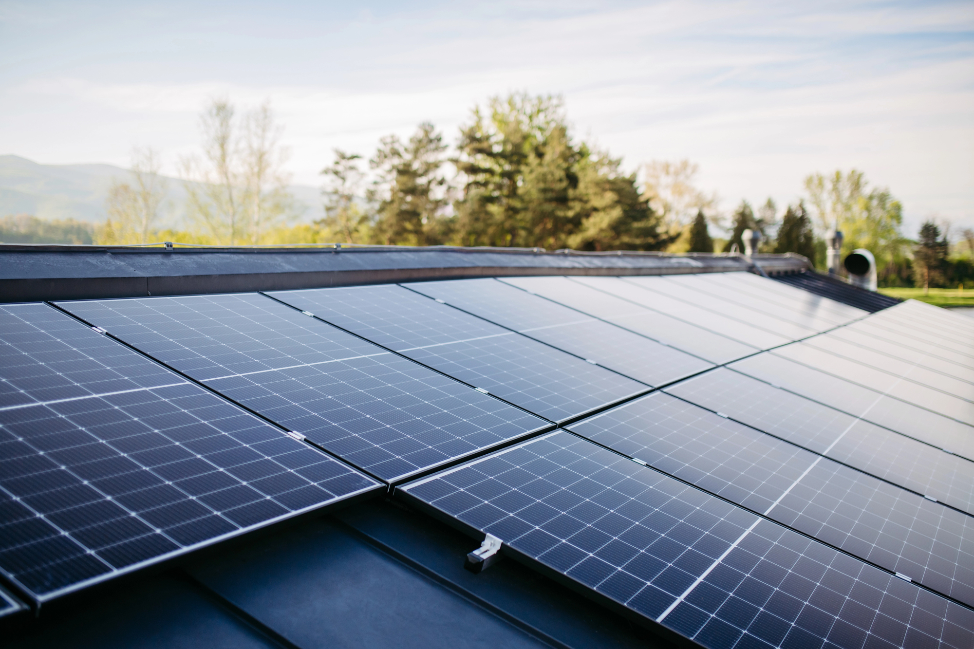 Rows of solar panels installed on a building roof with trees and a clear sky in the background.