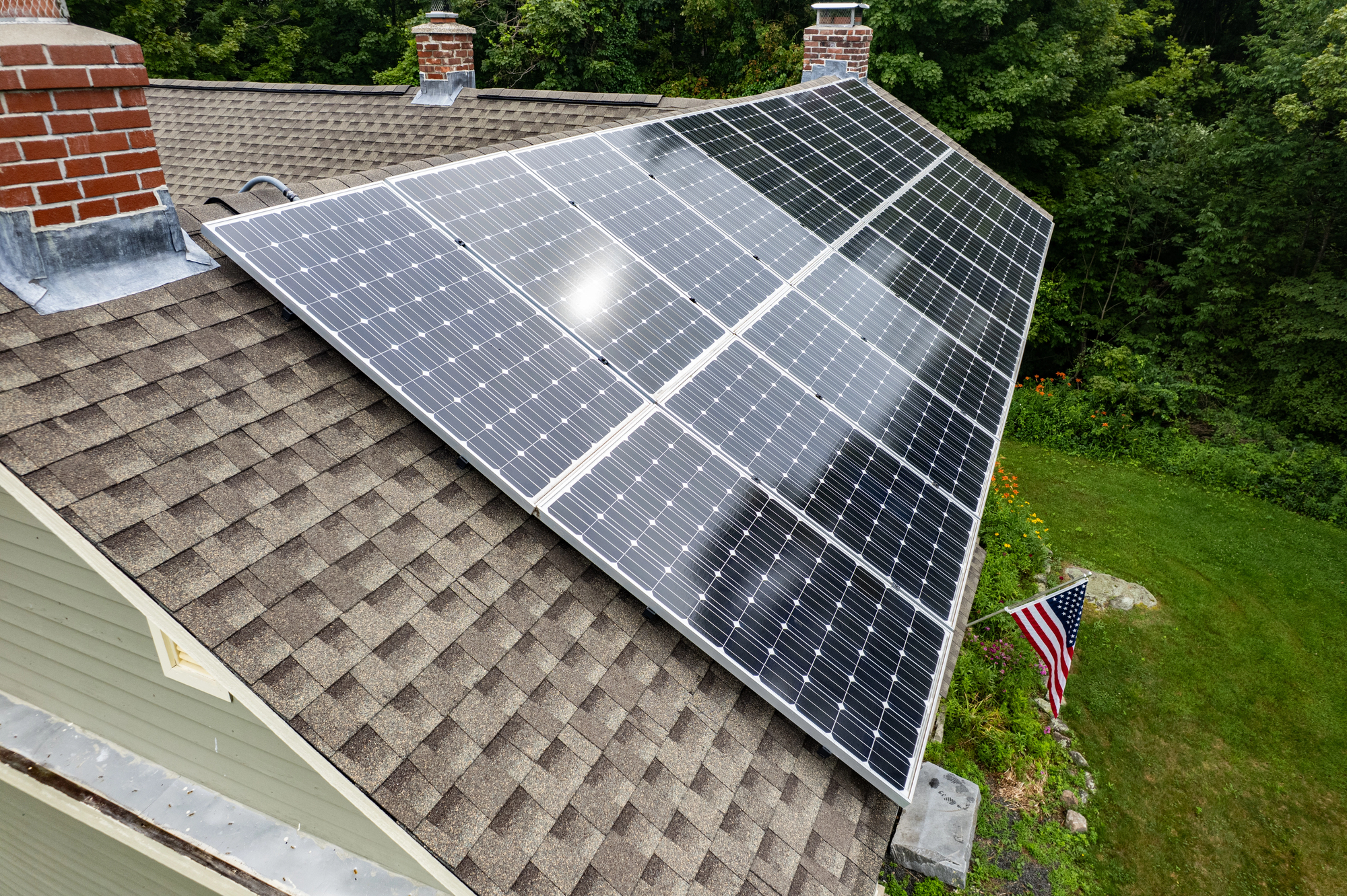 Solar panels installed on a sloped roof of a house with a shingle rooftop, surrounded by green trees and a lawn with an American flag.