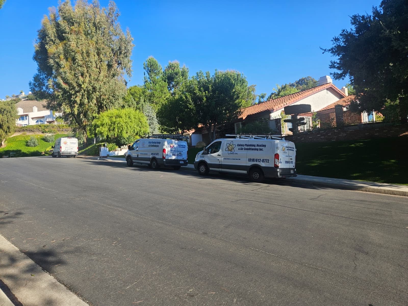 Three white service vans are parked on a suburban street with houses and trees on both sides under a clear blue sky.