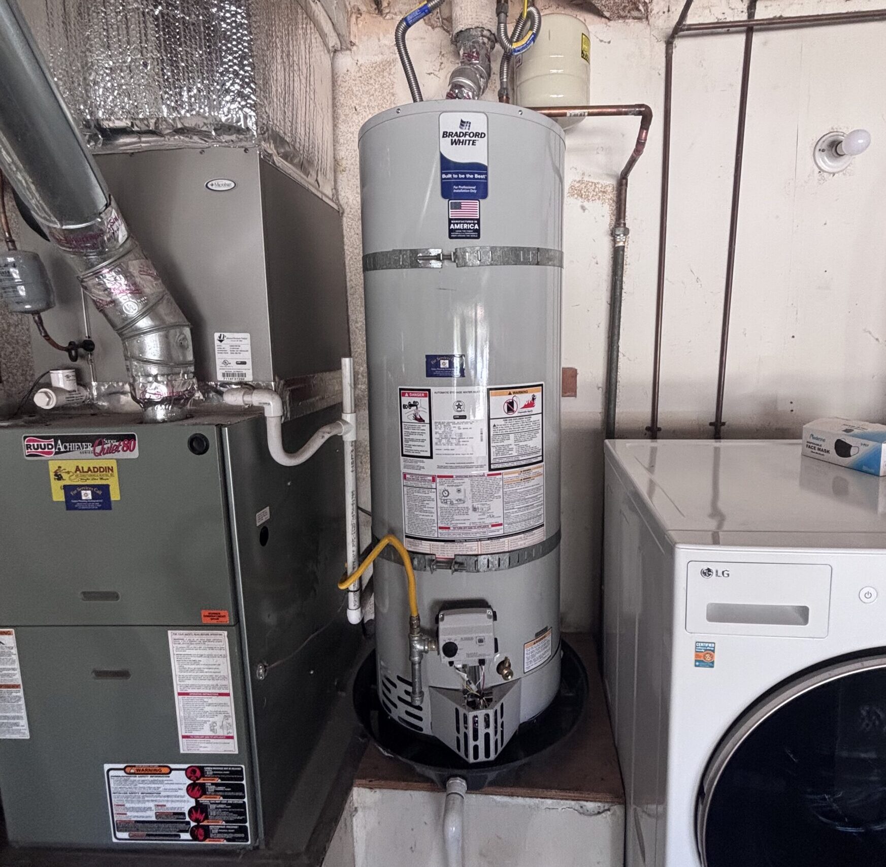 A water heater is installed next to a furnace and a front-load washing machine in a utility room with exposed pipes and wires.
