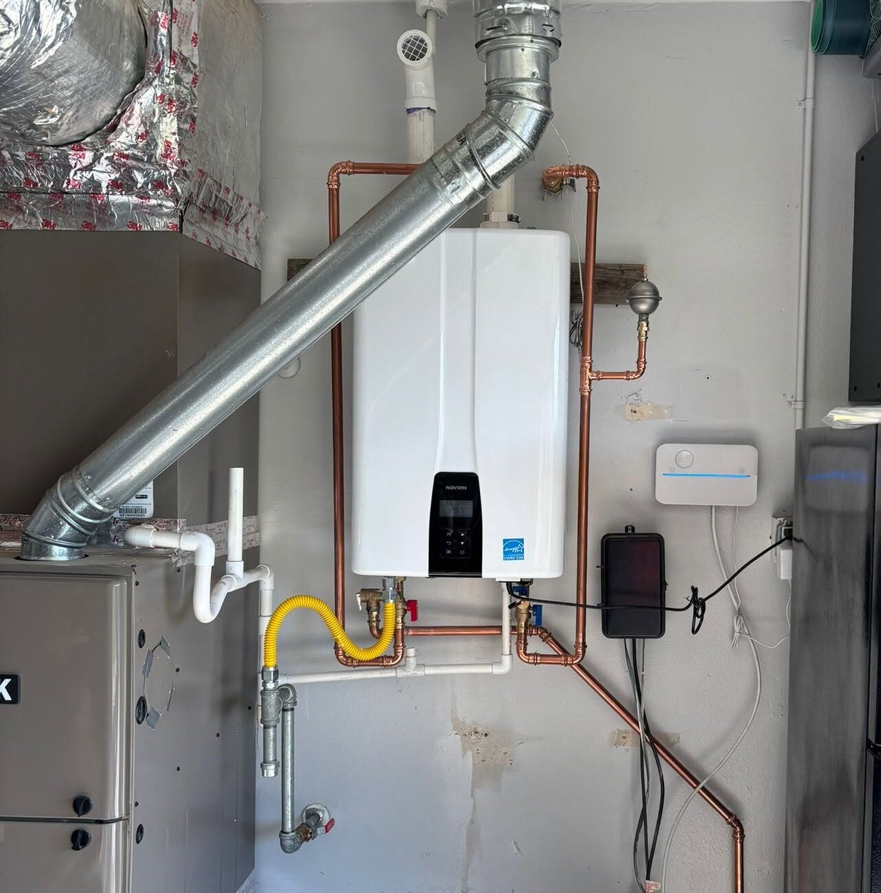 A tankless water heater is mounted on a garage wall, with visible pipes and ducts connected to nearby HVAC and utility equipment.