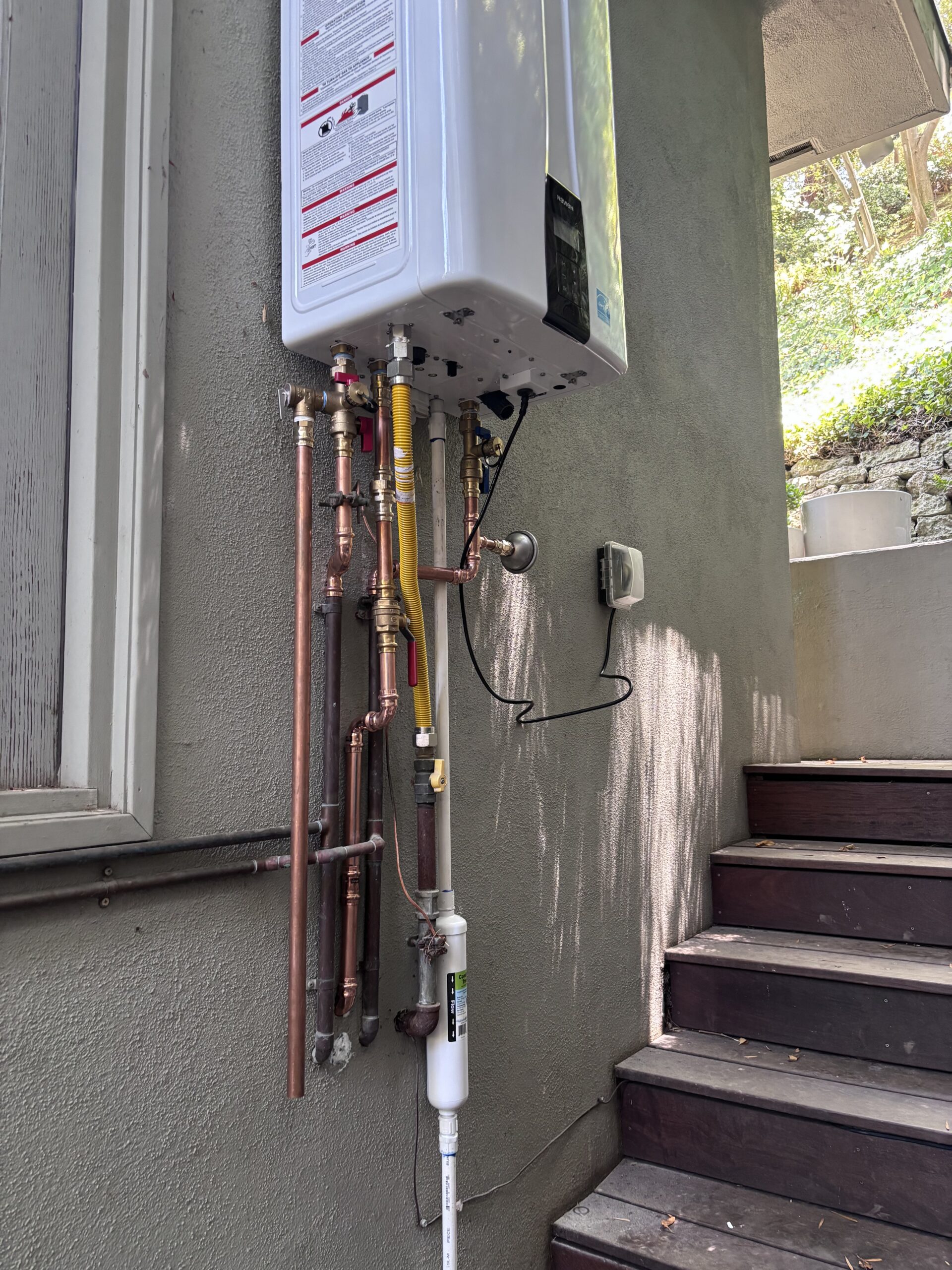 Outdoor tankless water heater mounted on a wall with exposed copper pipes, yellow gas line, wiring, and a filtration system visible next to a set of wooden stairs.
