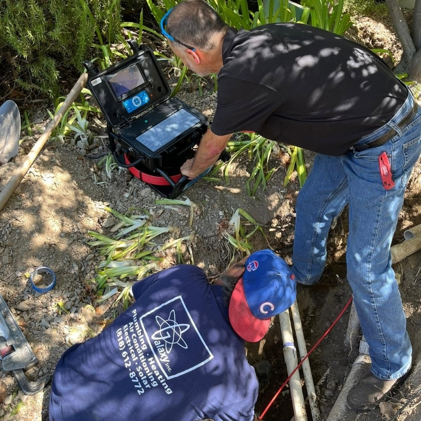 Two workers use electronic equipment to inspect underground pipes in an outdoor area with plants and soil.